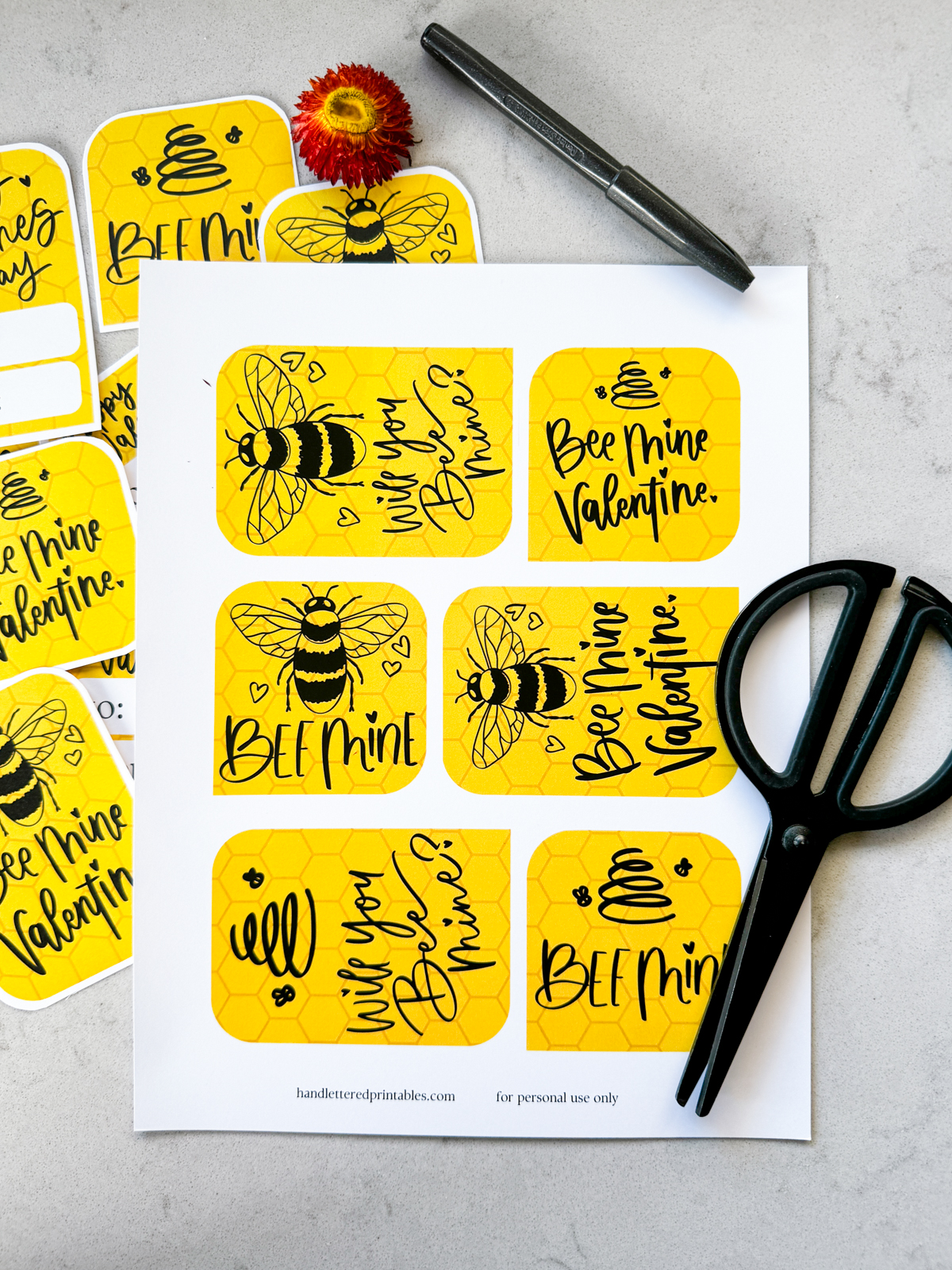 image of printed bee themed valentines cards on marble counter, all ready to be cut out. valentines cards read: bee mine, will you bee mine, and bee mine valentine with illustrations of a bee and of a line art bee hive. reverse has space for names and says 'happy valentines day'. all hand lettered in black ink with yellow honeycomb background