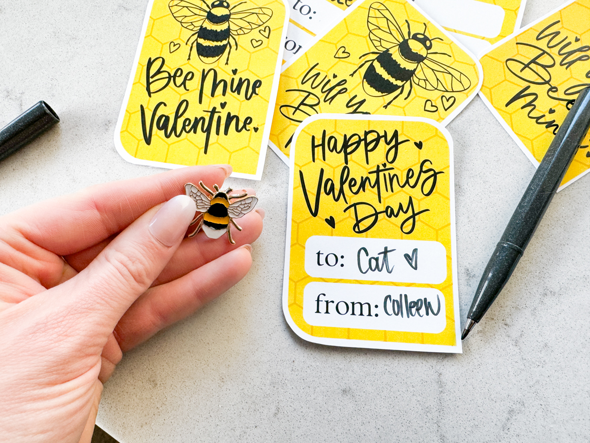 image of printed bee themed valentines cards on marble counter, cut to size and addressed with a black pen. hand holding an enamel bee pin to pair with the valentines cards
