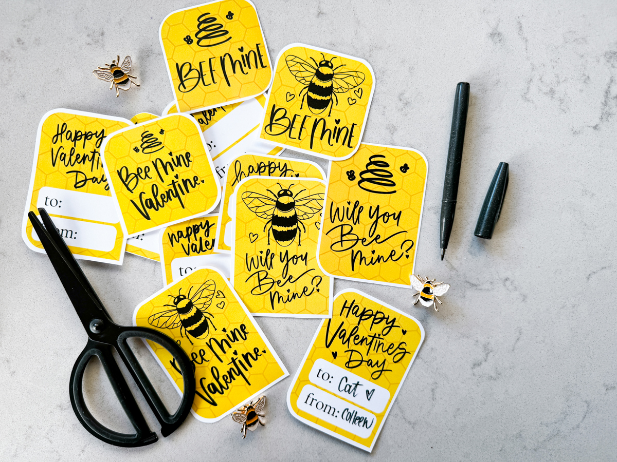 image of printed bee themed valentines cards on marble counter, already cut out. valentines cards read: bee mine, will you bee mine, and bee mine valentine with illustrations of a bee and of a line art bee hive. reverse has space for names and says 'happy valentines day'. all hand lettered in black ink with yellow honeycomb background