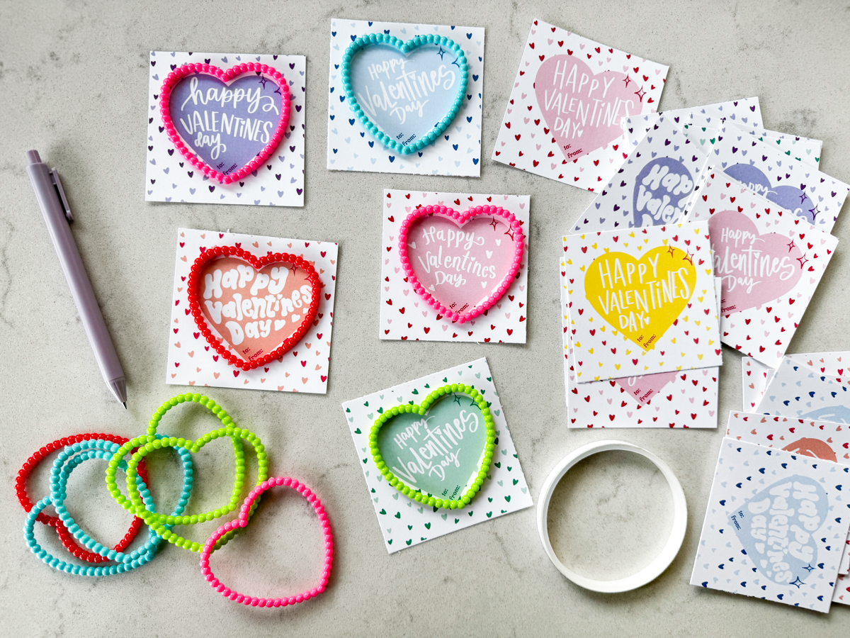 free printable happy valentines day cards in various candy colors shown cut to size (square) with heart bracelets on marble countertop