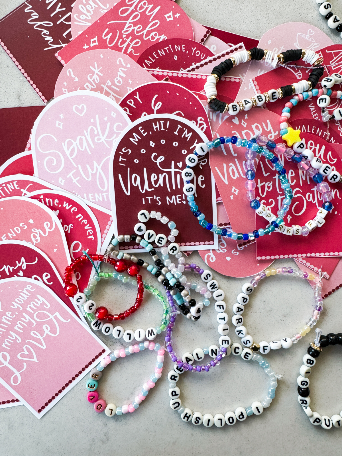 taylor swift themed valentines day cards printed and cut to size (pink and deep burgundy color with white hand lettered song lyrics) shown on marble countertop with beaded friendship bracelets