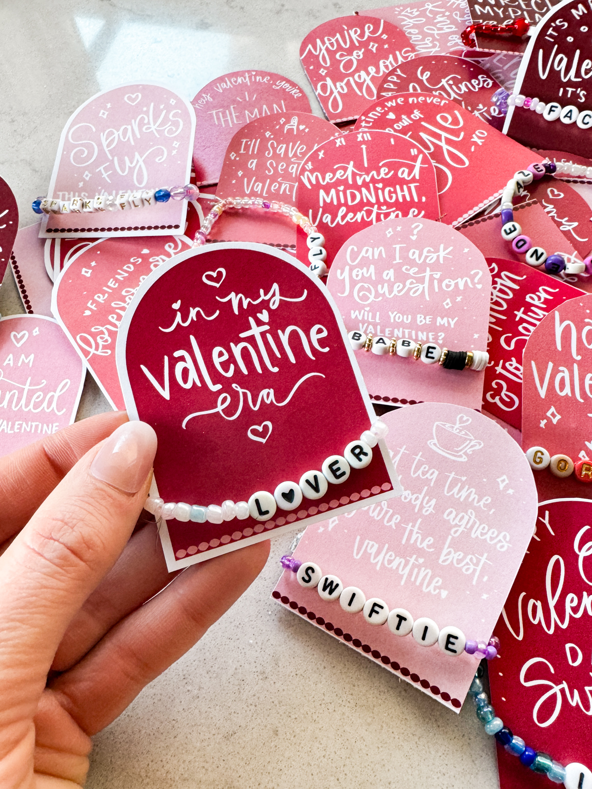 taylor swift themed valentines day cards printed and cut to size (pink and deep burgundy color with white hand lettered song lyrics) shown on marble countertop with beaded friendship bracelets around the arched cards. shown valentine reads 'in my valentine era' with a 'lover bracelet'