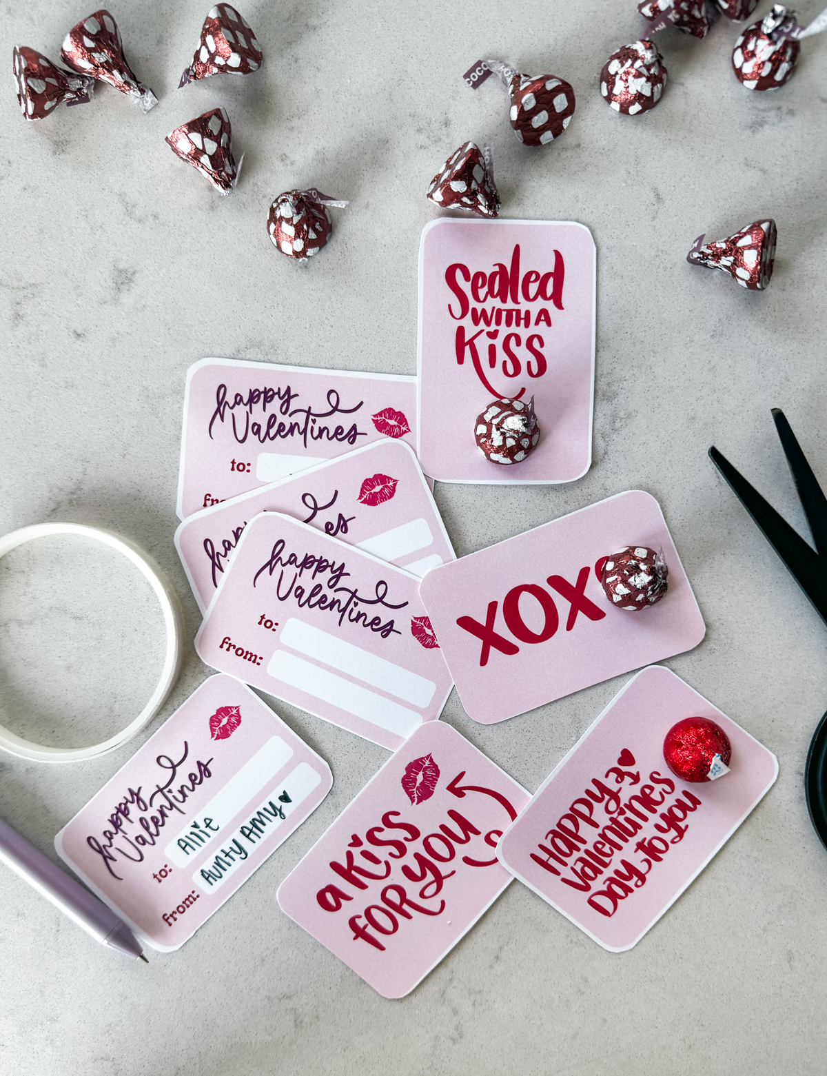 free printable valentines cards with pink backgrounds and red hand lettering, styled on marble countertop with black scissors, hershey's kisses, a purple pen and double sided tape