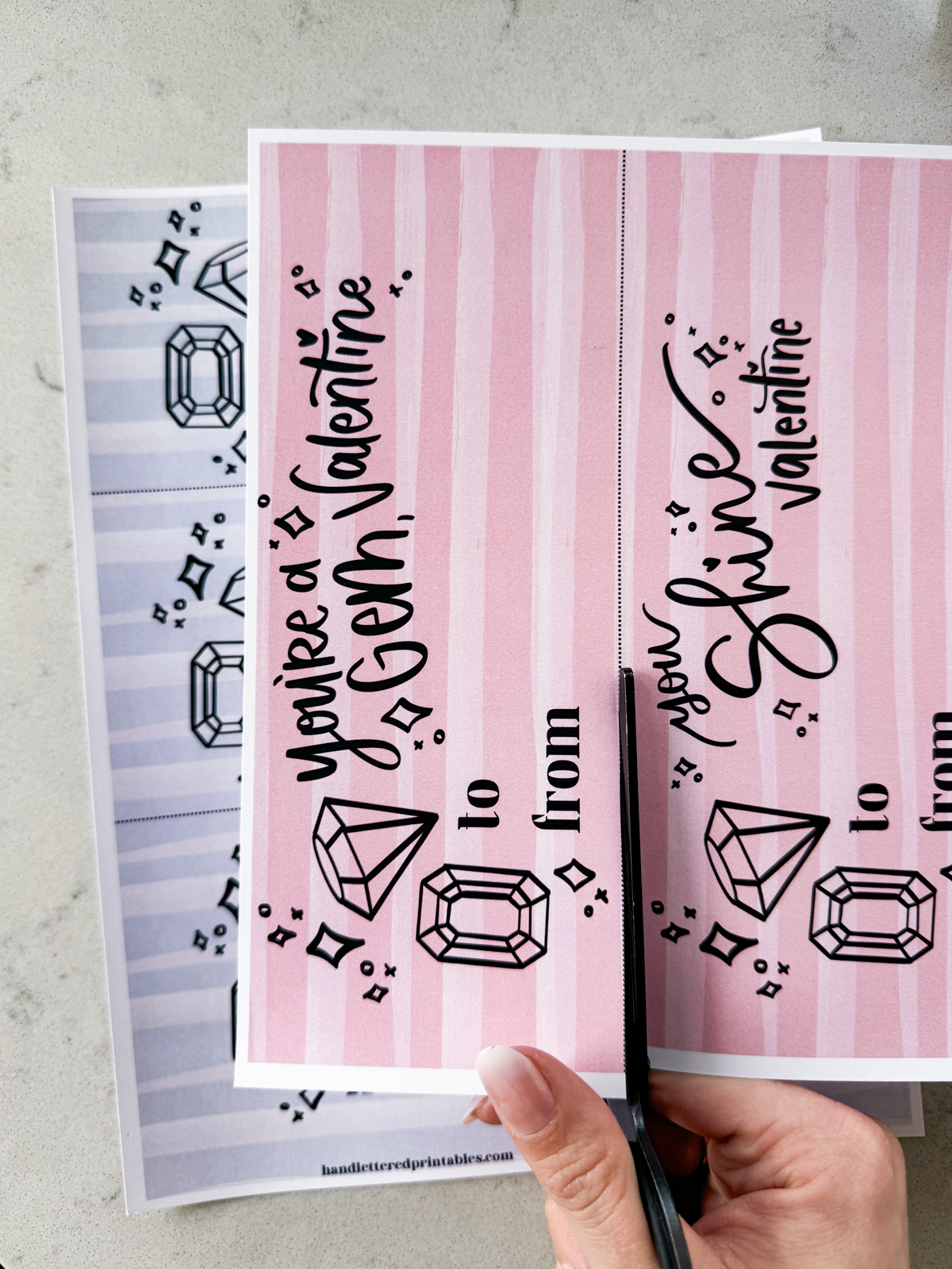 image of printed valentines with a gem theme, designed to pair with ring pops. printed and being cut to size on a marble counter shown hand lettered valentines in both pink and purple read:' you're a gem, valentine', 'you shine, valentine', and 'have a poppin' valentine's day'