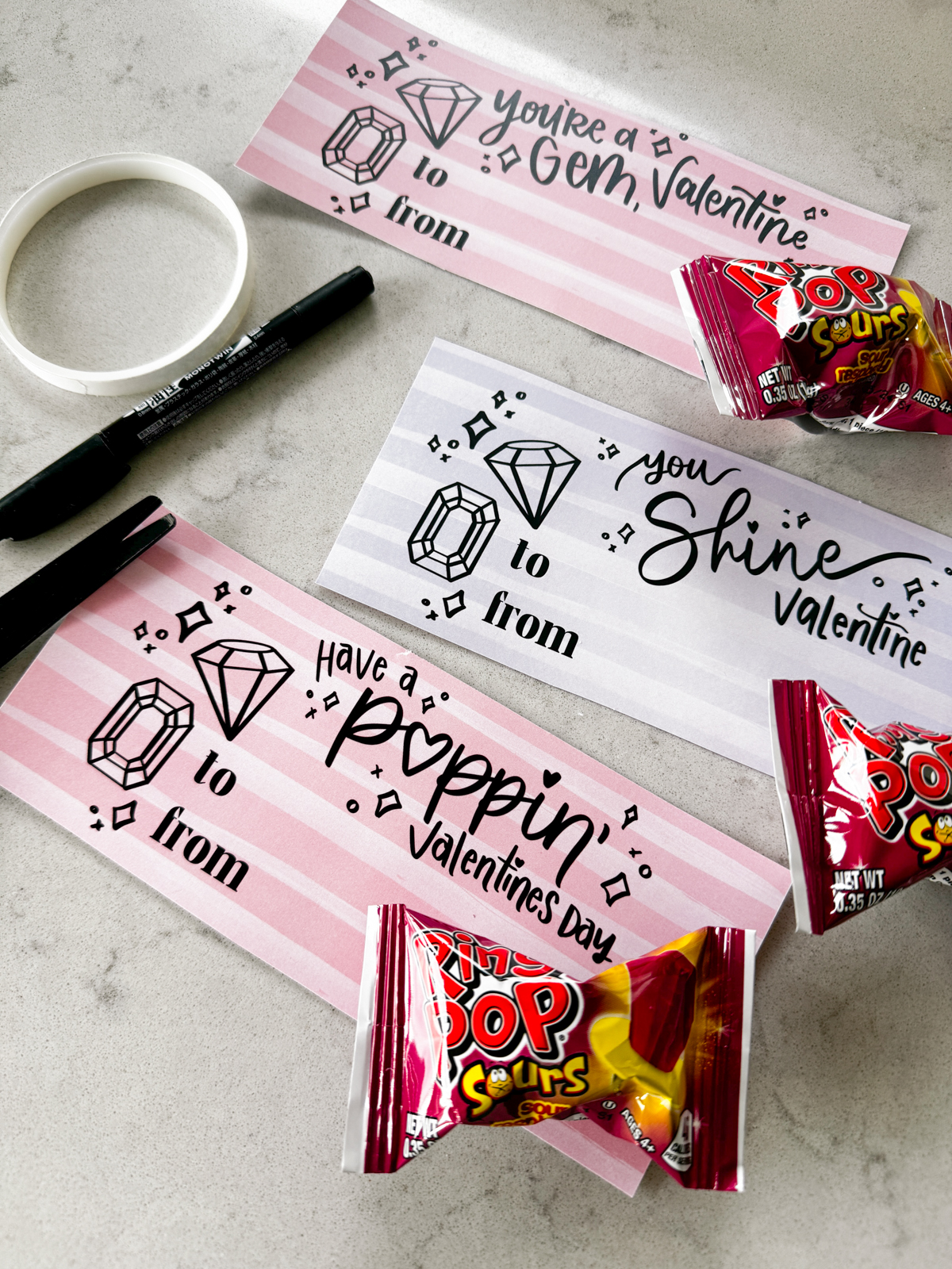 image of printed valentines with a gem theme, designed to pair with ring pops. printed and cut to size on a marble counter shown with ring pops in packaging taped to each valentine card with double sided tape hand lettered valentines in both pink and purple read:' you're a gem, valentine', 'you shine, valentine', and 'have a poppin' valentine's day'