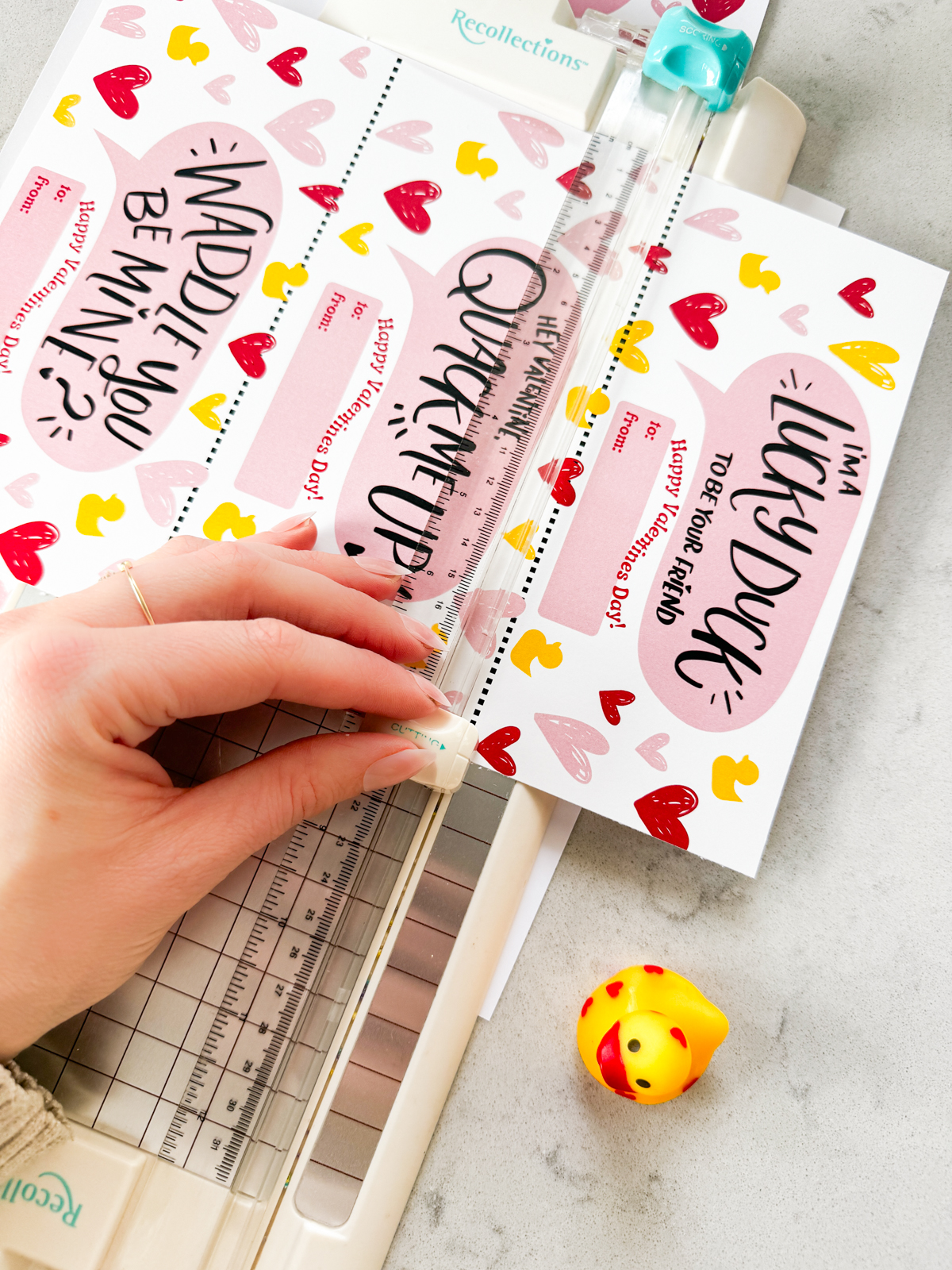 duck themed printable valentines day cards. cards shown read: waddle you be mine, hey valentine you quack me up and lucky to be your friend- all with a small happy valentines day. shown with rubber duckies, being cut to size with a paper cutter