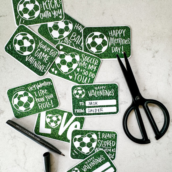 image shows soccer themed valentines cut to size with rounded corners on a marble countertop and styled with a black pen and black scissors valentines have a heart shaped soccer ball and a soccer ball with a heart on it and a grass background. valentines puns and sentiments include: happy valentines day, have a ball this valentines, hey valentine i like how you roll, soccer has my heart and so do you, i get a kick outta you, love (with a heart soccerball as the o), you've got game valentine, and i really scored with you as a friend reverse reads 'happy valentines' with room for to and from names. soccer/ american football