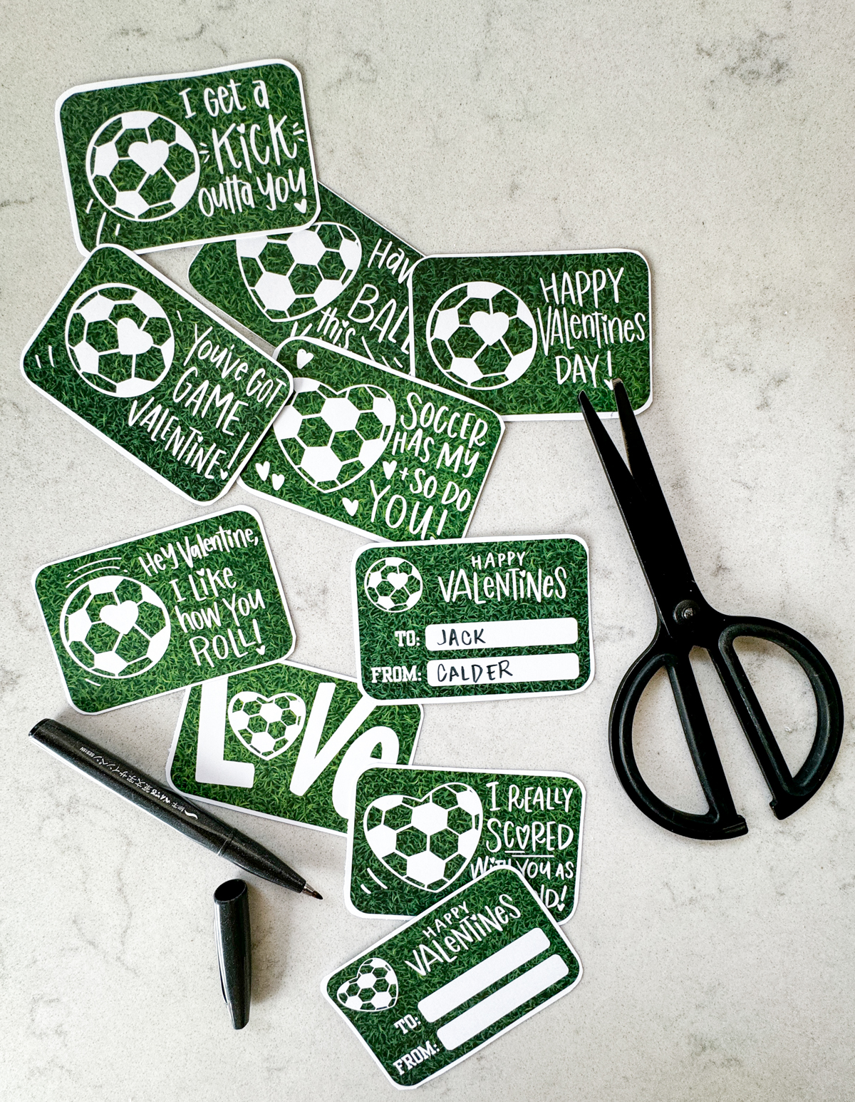 image shows soccer themed valentines cut to size with rounded corners on a marble countertop and styled with a black pen and black scissors valentines have a heart shaped soccer ball and a soccer ball with a heart on it and a grass background. valentines puns and sentiments include: happy valentines day, have a ball this valentines, hey valentine i like how you roll, soccer has my heart and so do you, i get a kick outta you, love (with a heart soccerball as the o), you've got game valentine, and i really scored with you as a friend reverse reads 'happy valentines' with room for to and from names. soccer/ american football
