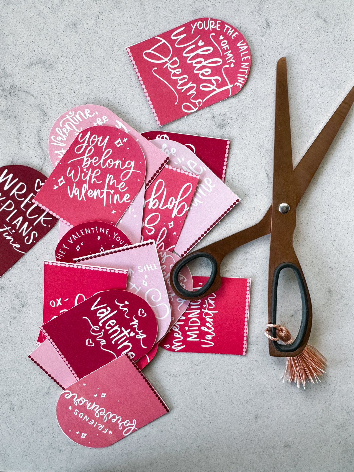 taylor swift themed valentines day cards printed and cut to size (pink and deep burgundy color with white hand lettered song lyrics) shown on marble countertop with rose gold scissors. some valentines read: you're the valentine of my wildest dreams, you belong with me valentine, in my valentine era, friends forevermore