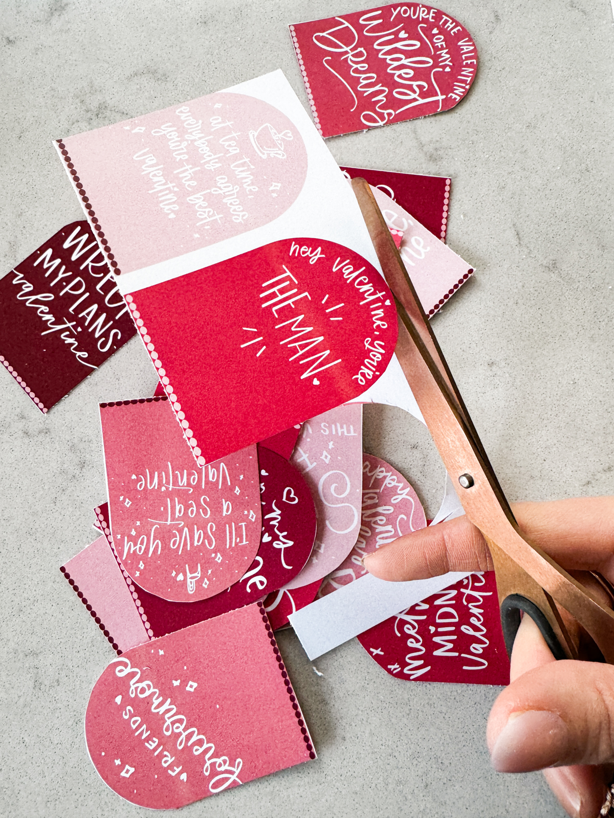 taylor swift themed valentines day cards printed and cut to size (arch shaped pink and deep burgundy color with white hand lettered song lyrics) shown on marble countertop being cut to size with rose gold scissors. valentine in focus reads: hey valentine, you're the man