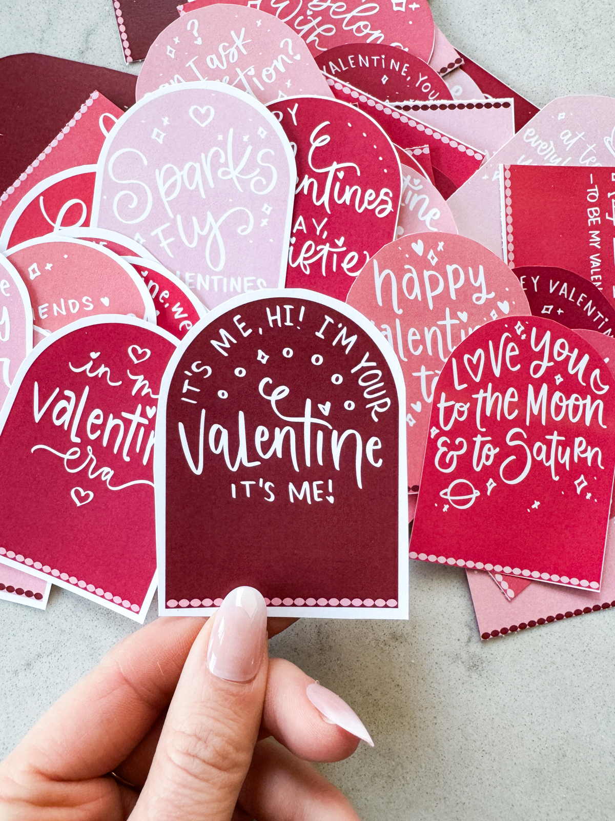 taylor swift themed valentines day cards printed and cut to size (arch shaped pink and deep burgundy color with white hand lettered song lyrics) shown on marble countertop. held valentine reads: it's me, hi! i'm your valentine, it's me!