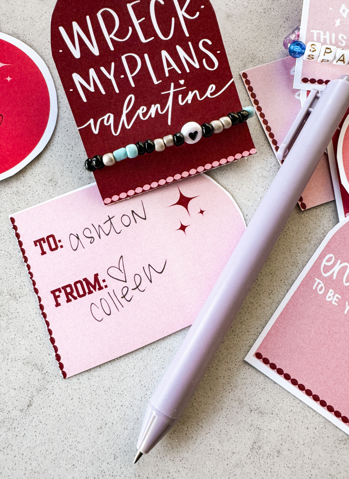image shows printable taylor swift valentines cards. one card is shown face up with the hand lettering 'wreck my plans, valentine' from taylor swift lyrics. valentine has a heart friendship bracelet second valentine shoes reverse with space for to and from both valentines are arch shape in shades of pink and red