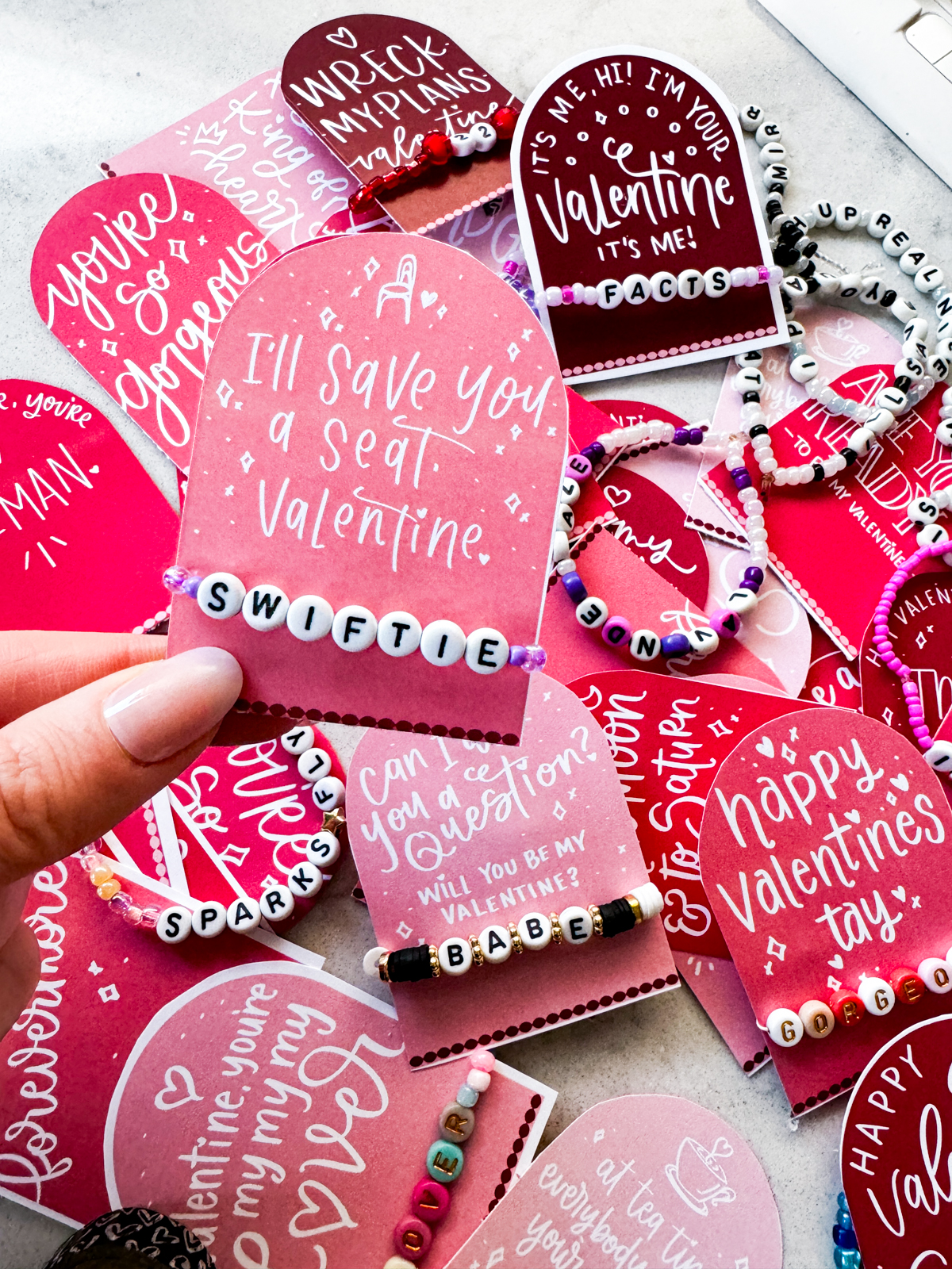 taylor swift themed valentines day cards printed and cut to size (pink and deep burgundy color with white hand lettered song lyrics) shown on marble countertop with beaded friendship bracelets. held valentine reads: i'll save you a seat, valentine with a 'swiftie' bracelet.