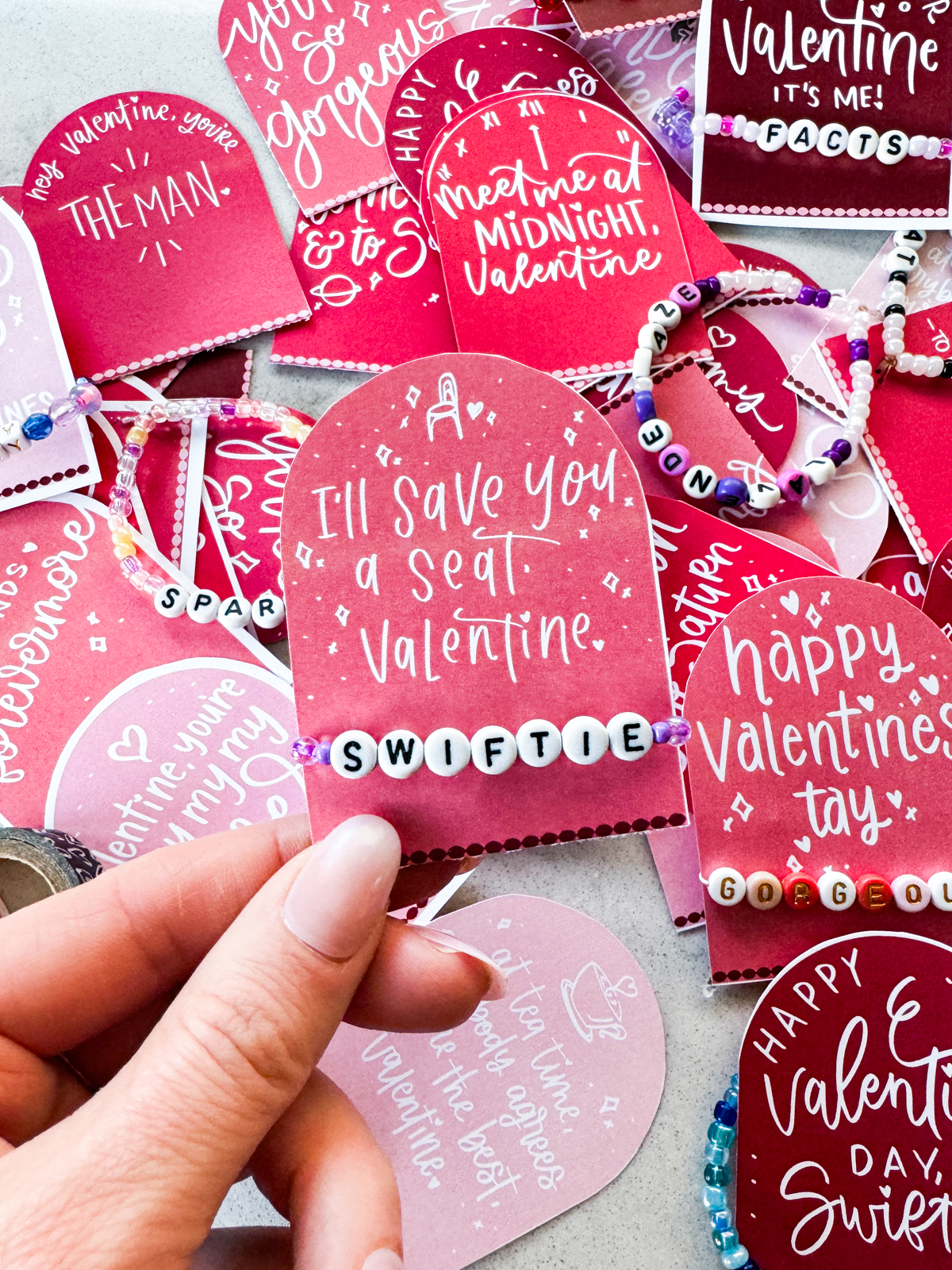 taylor swift themed valentines day cards printed and cut to size (pink and deep burgundy color with white hand lettered song lyrics) shown on marble countertop with beaded friendship bracelets. held valentine reads: i'll save you a seat, valentine with a 'swiftie' bracelet.