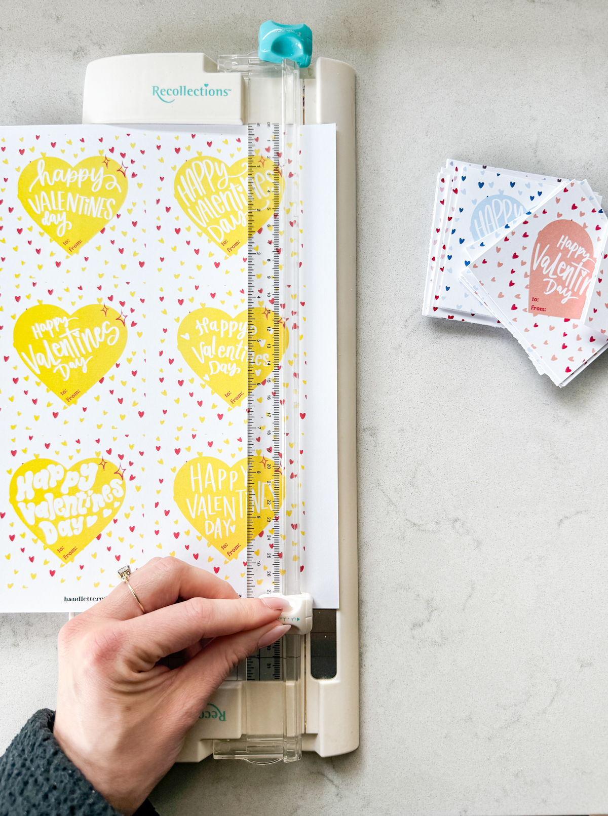 image shows happy valentines day printable cards being cut to size with a paper cutter. stacked valentines cut to size (pink, blue, orange) while yellow is being cut