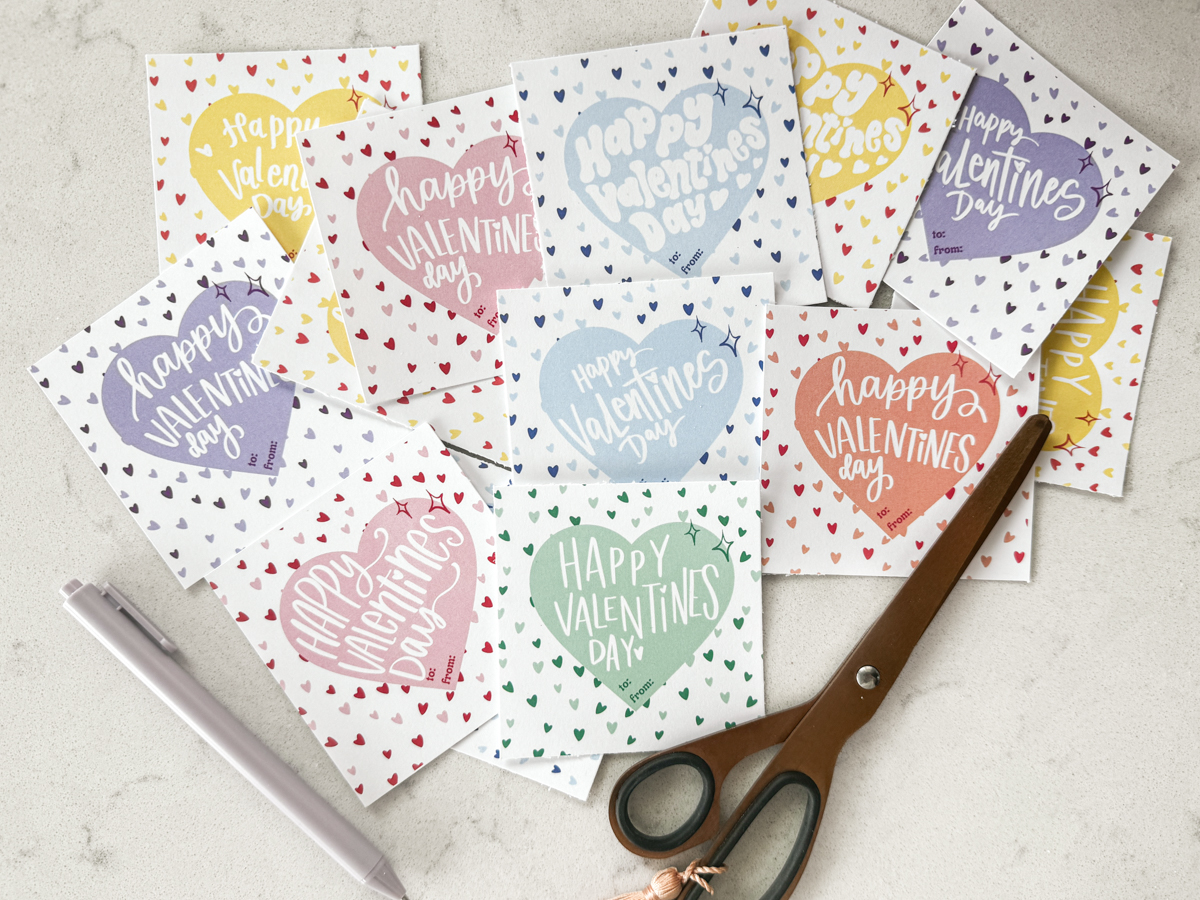 free printable happy valentines day cards in 6 candy colors (pink, purple, yellow, blue, orange, and green) shown cut to size (square) styled on marble countertop with purple pen and rose gold scissors.