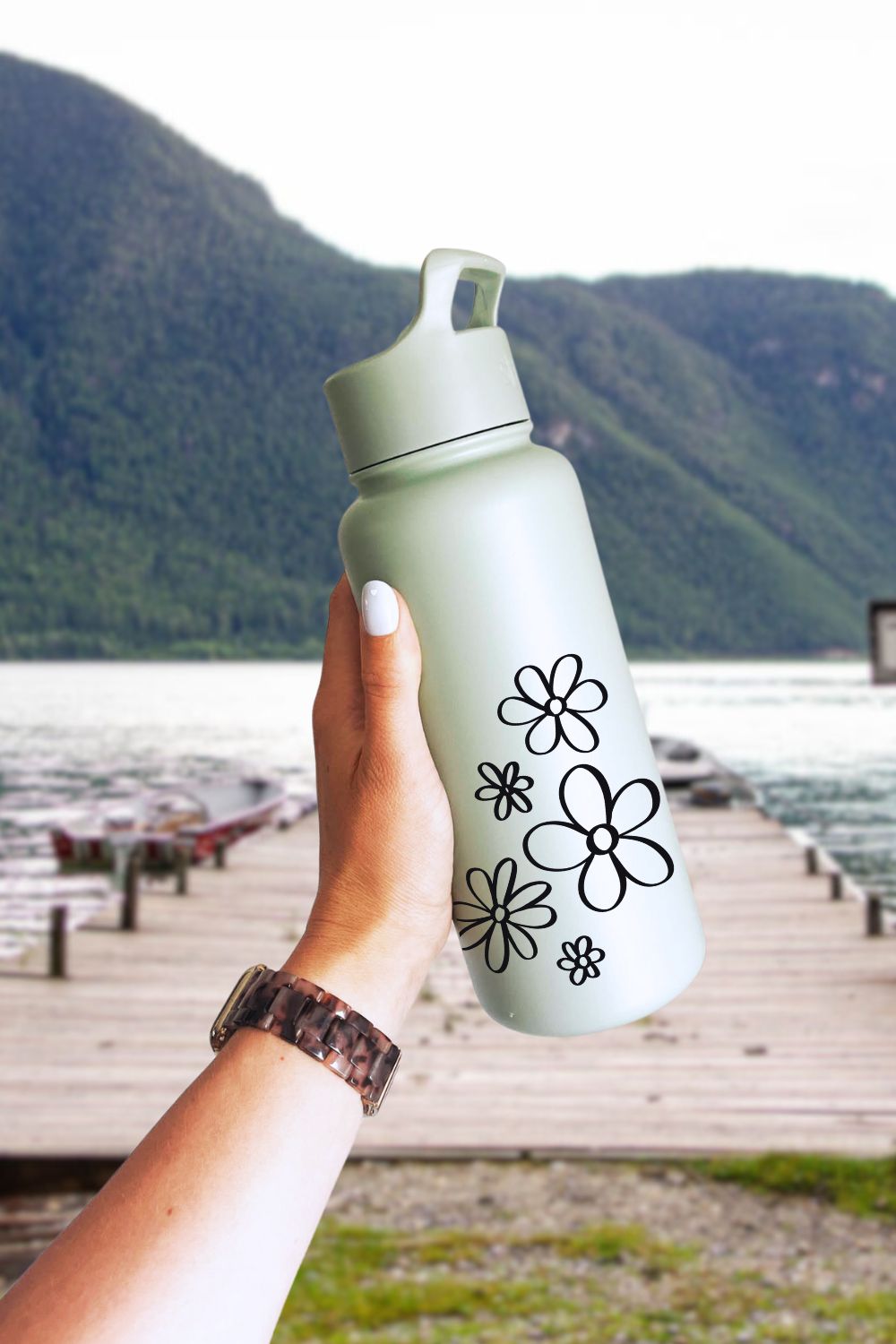 Hand drawn flowers design stuck onto a metal waterbottle held in front of a dock at a lake- free cut file cut with cricut of 5 flowers