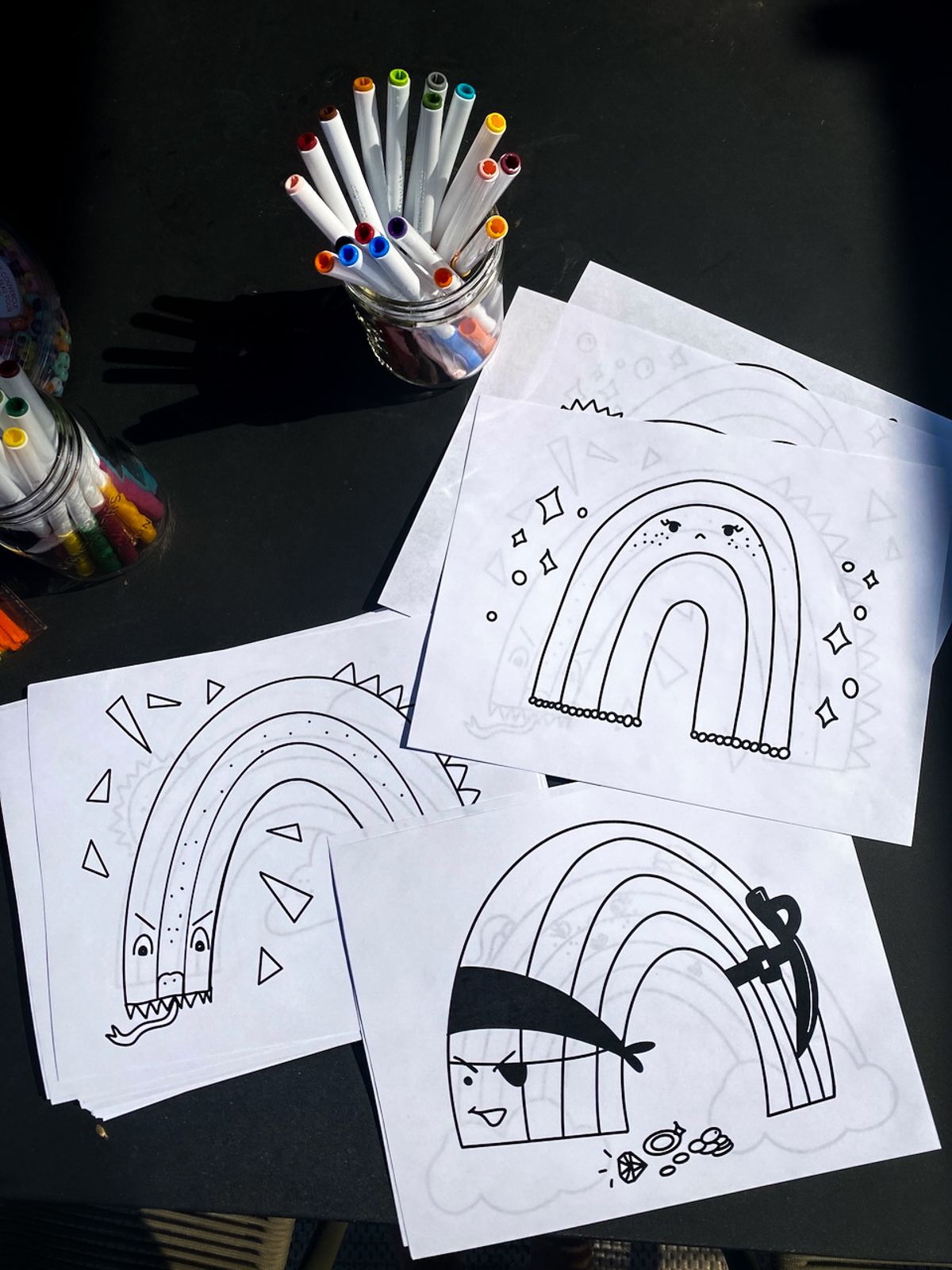 image shows free printable rainbow coloring sheets printed off and displayed on a table with markers in the sunshine