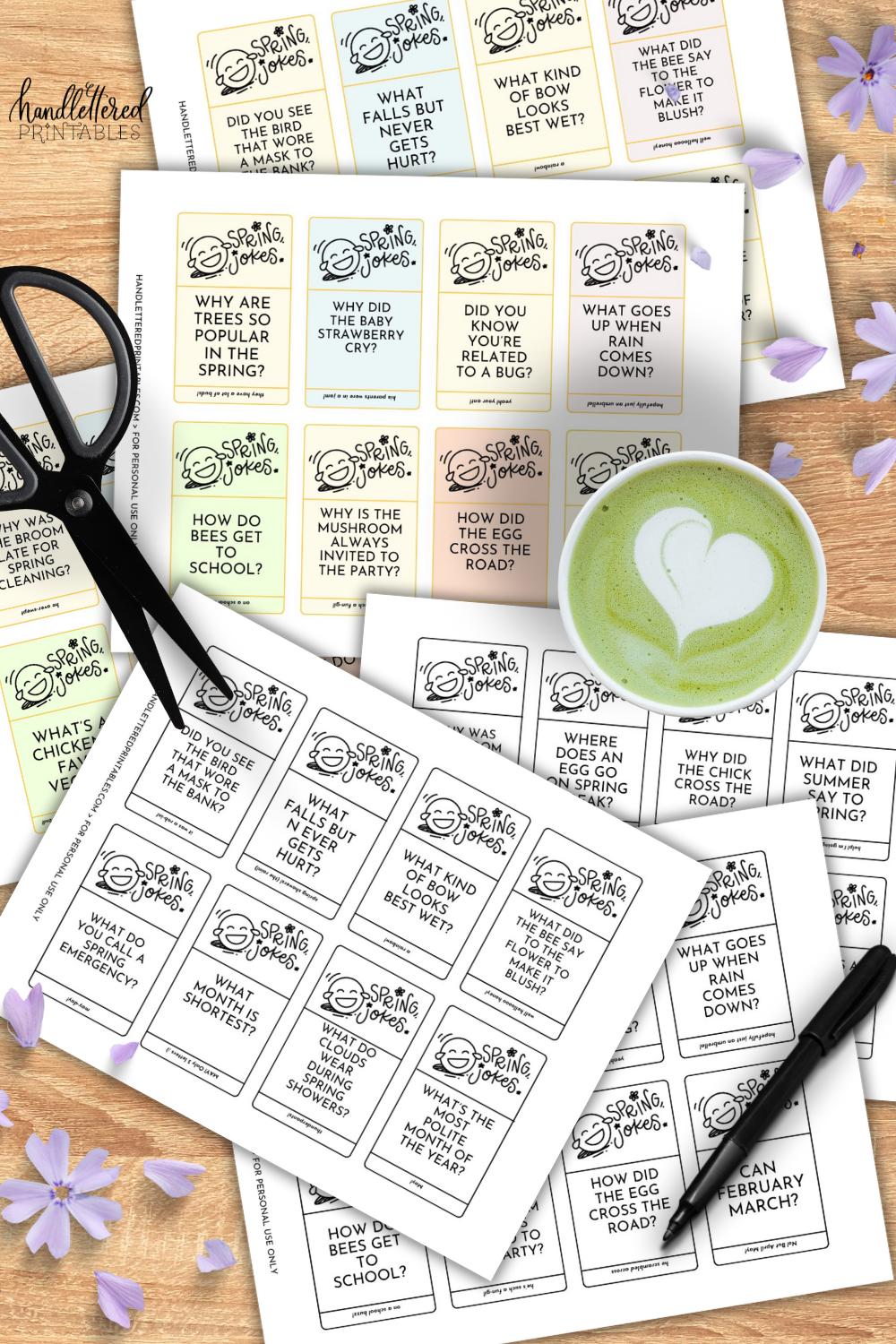 image of both color and black and white version of the spring jokes cards free printable printed on a wooden table with purple flowers, a matcha latte, pen and scissors. joke cards feature a hand lettered title and illustrated laughing emoji with the jokes.