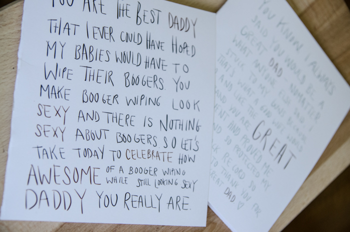 two free printable father's day cards on table, top card reads: image of funny fathers day card: woman's hand holding a card in a messy living room with baby in the background. card reads: you are the best daddy that I could ever have hoped my babies would have to wipe their boogers. You make booger wiping look sexy. And there is nothing sexy about boogers. So let's take today to celebrate how awesome of a booger wiping while still looking sexy you really are.