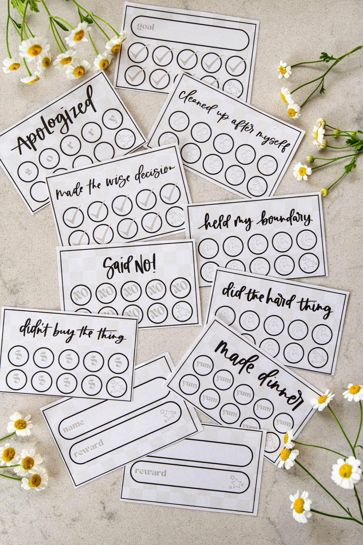 image of printed punch cards for adults, cut to size on countertop with cut flowers punch cards have a soft checkerboard background with hand lettered goals like 'said no!', 'didn't buy the thing', 'made dinner', 'held my boundary' and 'made the wise decision'