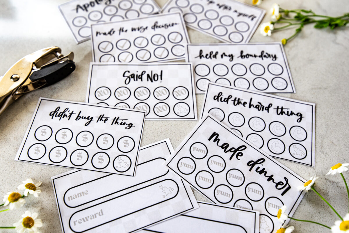 image of printed punch cards for adults, cut to size on countertop with hole punch and cut flowers punch cards have a soft checkerboard background with hand lettered goals like 'said no!', 'didn't buy the thing', 'made dinner', 'held my boundary' and 'made the wise decision'