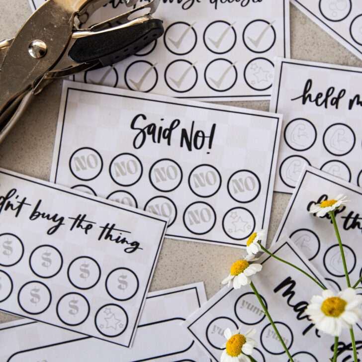 image of printed punch cards for adults, cut to size on countertop with hole punch and cut flowers punch cards have a soft checkerboard background with hand lettered goals like 'said no!', 'didn't buy the thing', 'made dinner', 'held my boundary' and 'made the wise decision'