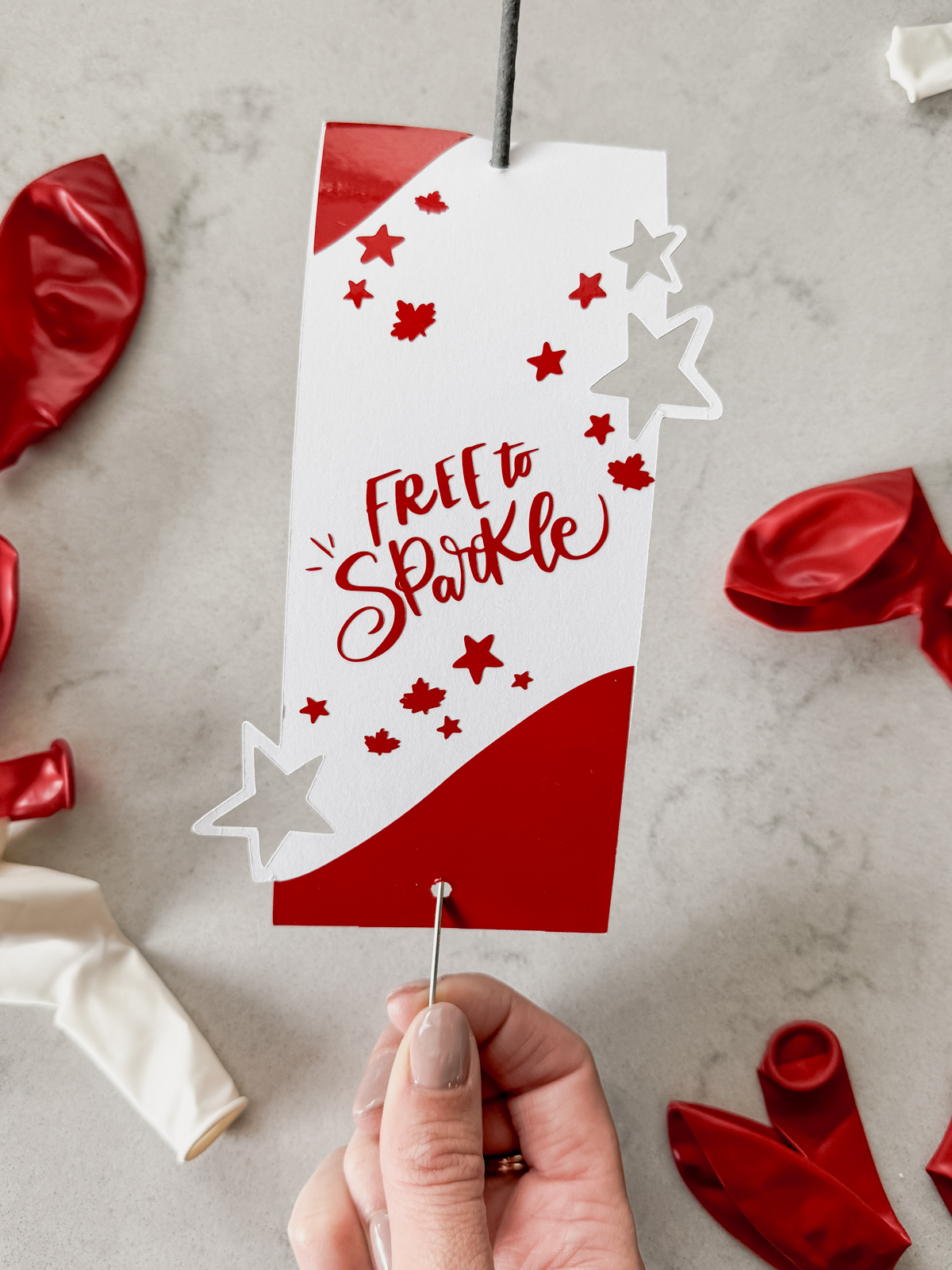 image of sparkler holder made with cricut- design has stars and maple leaves and the hand lettering 'free to sparkle' sparkler holder shown held over marble countertop with red and white balloons
