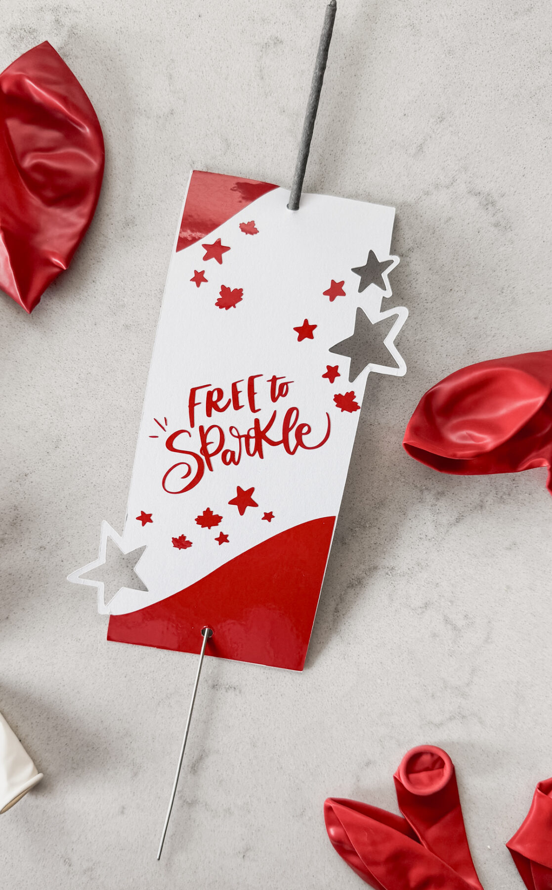 image of sparkler holder made with cricut- design has stars and maple leaves and the hand lettering 'free to sparkle' sparkler holder shown on marble countertop with red and white balloons