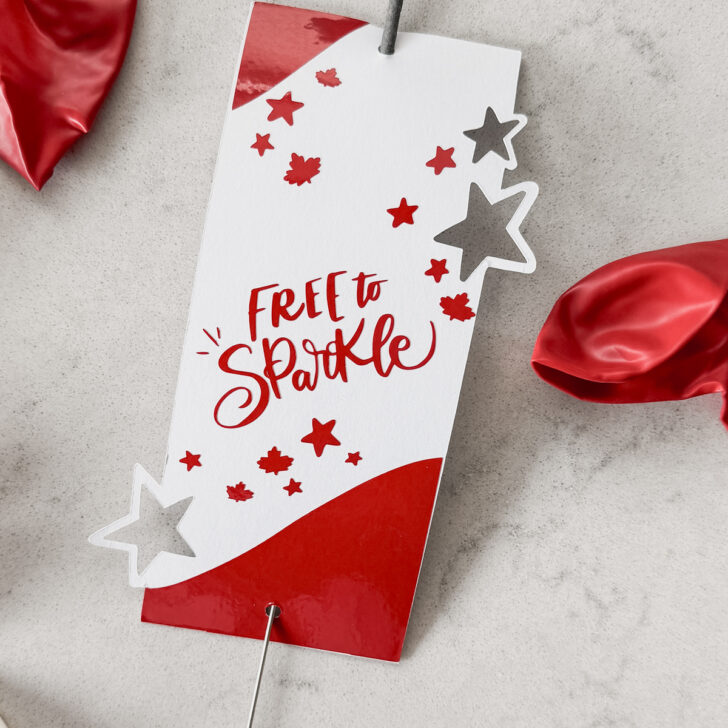 image of sparkler holder made with cricut- design has stars and maple leaves and the hand lettering 'free to sparkle' sparkler holder shown on marble countertop with red and white balloons
